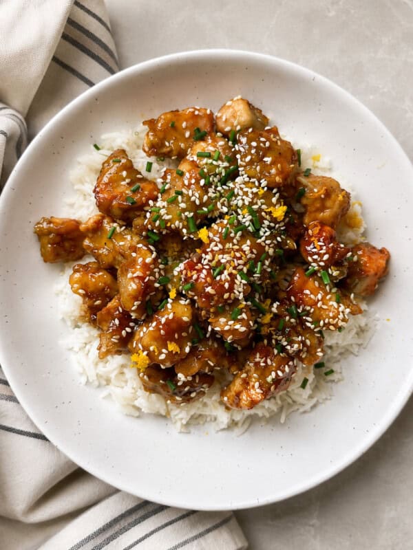 A white plate of rice topped with saucy, golden-brown chicken pieces from an easy orange chicken recipe, garnished with sesame seeds and chopped herbs, sits on a light-colored surface next to a striped cloth napkin.