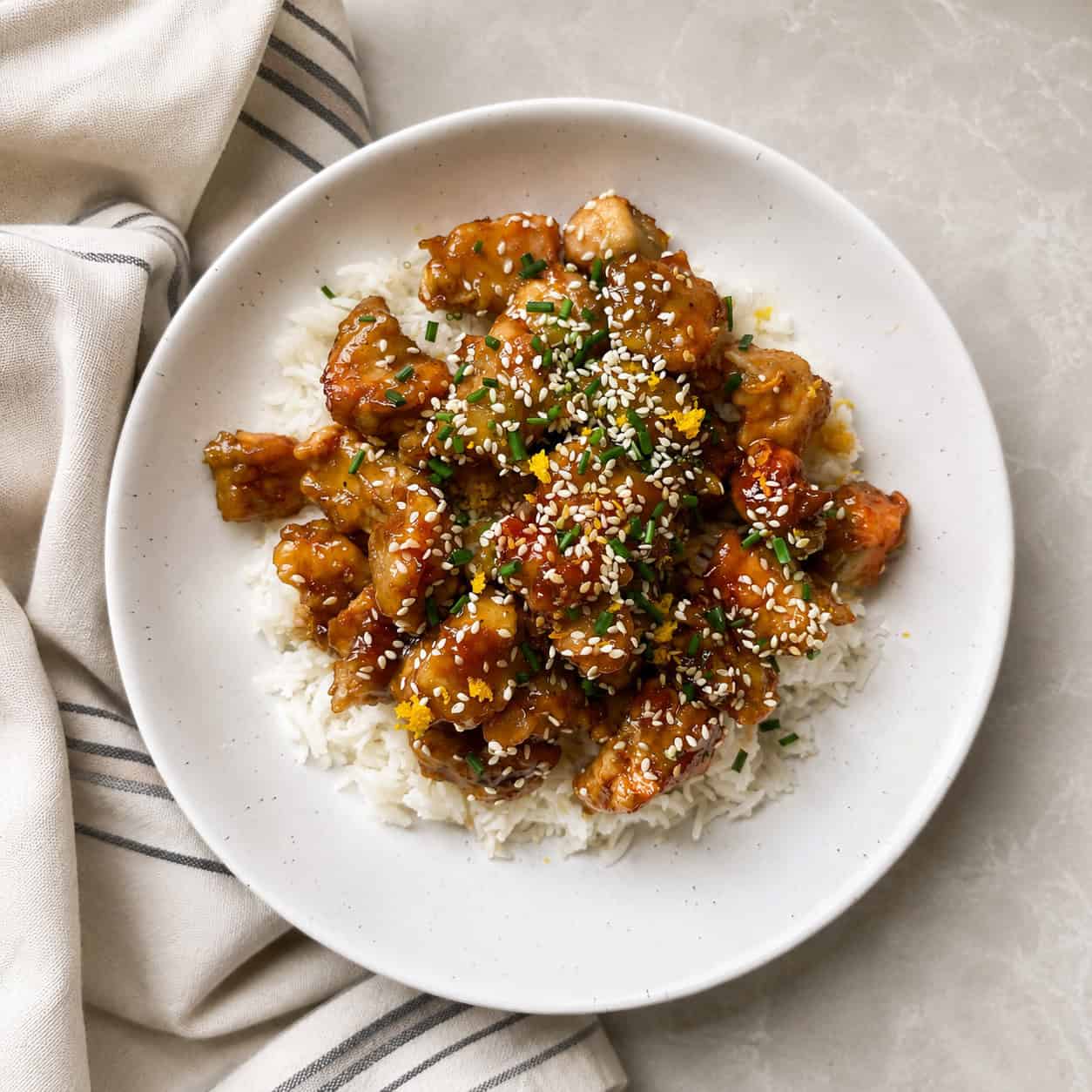 A white plate of rice topped with saucy, golden-brown chicken pieces from an easy orange chicken recipe, garnished with sesame seeds and chopped herbs, sits on a light-colored surface next to a striped cloth napkin.