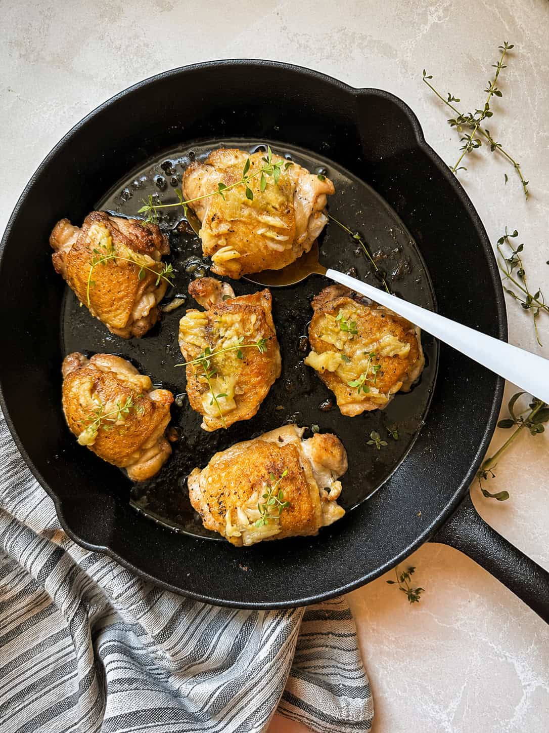 Six golden-brown chicken thighs garnished with herbs in a black cast iron skillet. A white spatula rests beside the chicken, and fresh thyme sprigs and a striped towel are nearby.