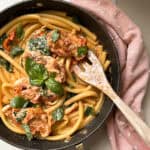 A skillet with pasta and sliced chicken in a creamy sauce, topped with fresh basil leaves. A wooden spoon rests in the pan. The skillet is placed on a light surface with a pink polka dot cloth beside it.