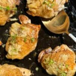 Golden brown chicken thighs garnished with herbs and diced garlic, sizzling in a black skillet with a golden spoon beside them.