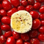A round slice of herbed goat cheese drizzled with olive oil sits on a bed of seasoned cherry tomatoes, evoking the flavors of Creamy Boursin & Tomato Pasta.