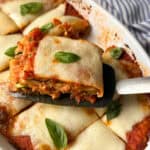 A close-up of a baked dish featuring layered pieces of eggplant parmesan topped with melted cheese and garnished with fresh basil leaves. A slice is being lifted with a spatula from the white baking dish. A striped cloth is visible in the background.