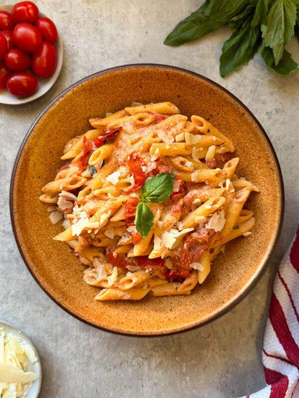 A bowl of Creamy Boursin & Tomato Pasta with tomato sauce, garnished with basil and grated cheese. Surrounding the bowl are a plate of cherry tomatoes, extra grated cheese, fresh basil, and a red striped towel on a light surface.