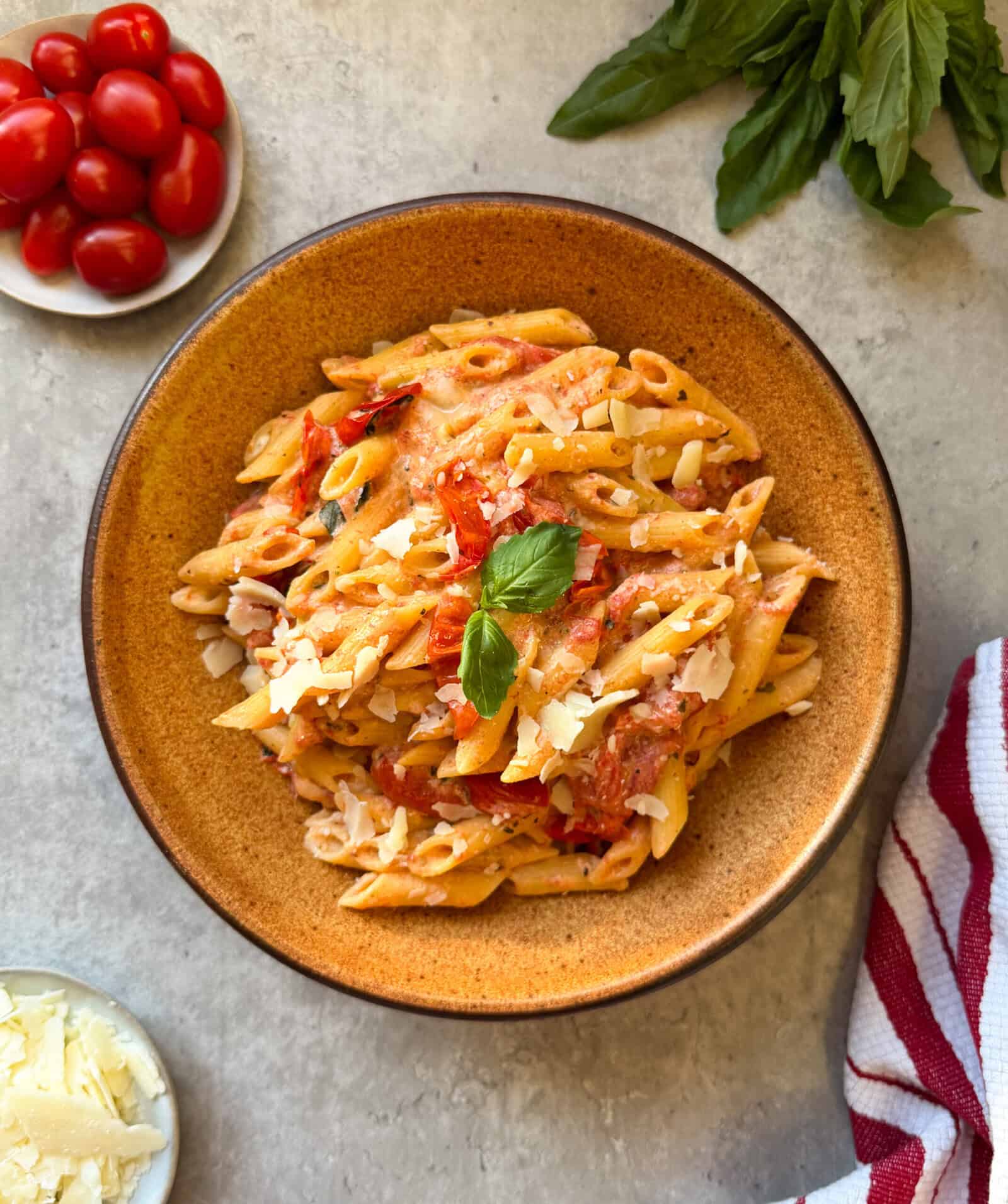 A bowl of Creamy Boursin & Tomato Pasta with tomato sauce, garnished with basil and grated cheese. Surrounding the bowl are a plate of cherry tomatoes, extra grated cheese, fresh basil, and a red striped towel on a light surface.