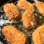 Golden brown croquettes frying in bubbling oil in a black pan. The croquettes have a crispy, breaded coating, with one showing a hint of melted filling.