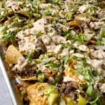 A tray of loaded nachos topped with melted cheese, seasoned ground beef, diced pickles, shredded lettuce, sesame seeds, and a drizzle of special sauce.