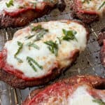 Close-up of several pieces of chicken parmesan on a wire rack. Each piece is topped with melted mozzarella cheese, tomato sauce, and garnished with sliced fresh basil. The golden-brown crust is visible under the sauce and cheese.