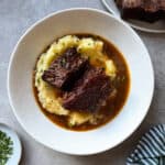 A white bowl filled with mashed potatoes, topped with two pieces of braised beef short ribs in brown gravy and garnished with chopped herbs, sits on a gray surface beside a green striped cloth and a small plate of chopped herbs.