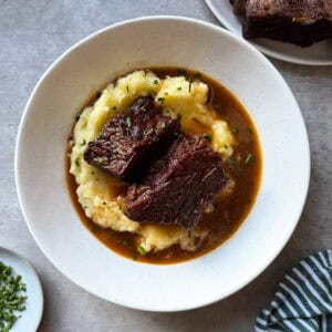 A white bowl filled with mashed potatoes, topped with two pieces of braised beef short ribs in brown gravy and garnished with chopped herbs, sits on a gray surface beside a green striped cloth and a small plate of chopped herbs.