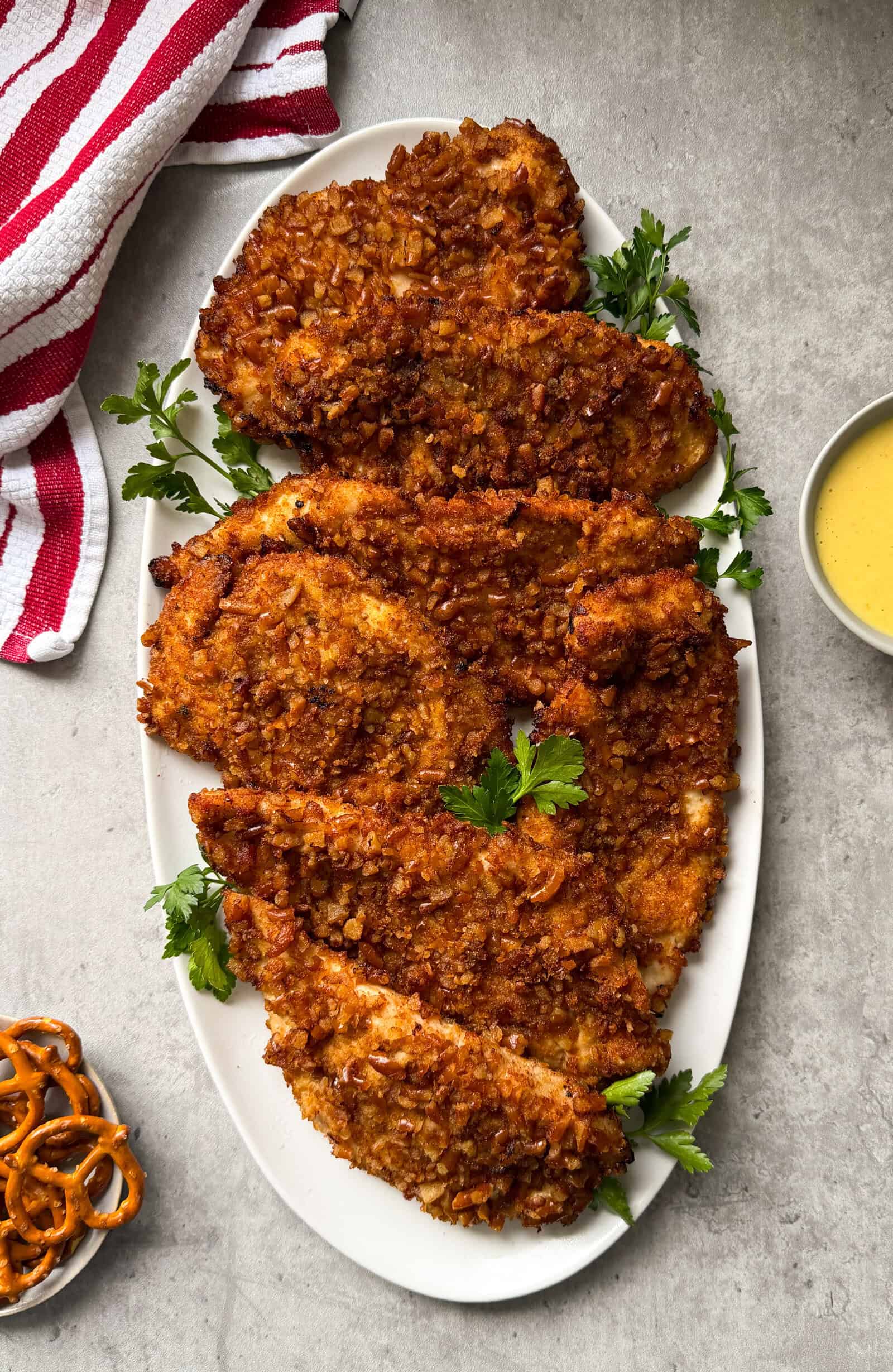 A white oval platter holds several pieces of crispy, golden-brown pretzel crusted chicken garnished with parsley, next to a yellow honey mustard dipping sauce, pretzels, and a red-and-white striped towel on a gray surface.
