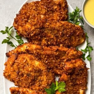 A white oval plate holds five crispy, golden-brown pretzel crusted chicken cutlets garnished with fresh parsley, with a small bowl of yellow dipping sauce beside them—an irresistible pretzel crusted chicken recipe.