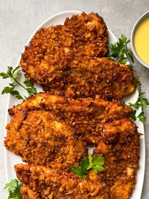 A white oval plate holds five crispy, golden-brown pretzel crusted chicken cutlets garnished with fresh parsley, with a small bowl of yellow dipping sauce beside themโan irresistible pretzel crusted chicken recipe.