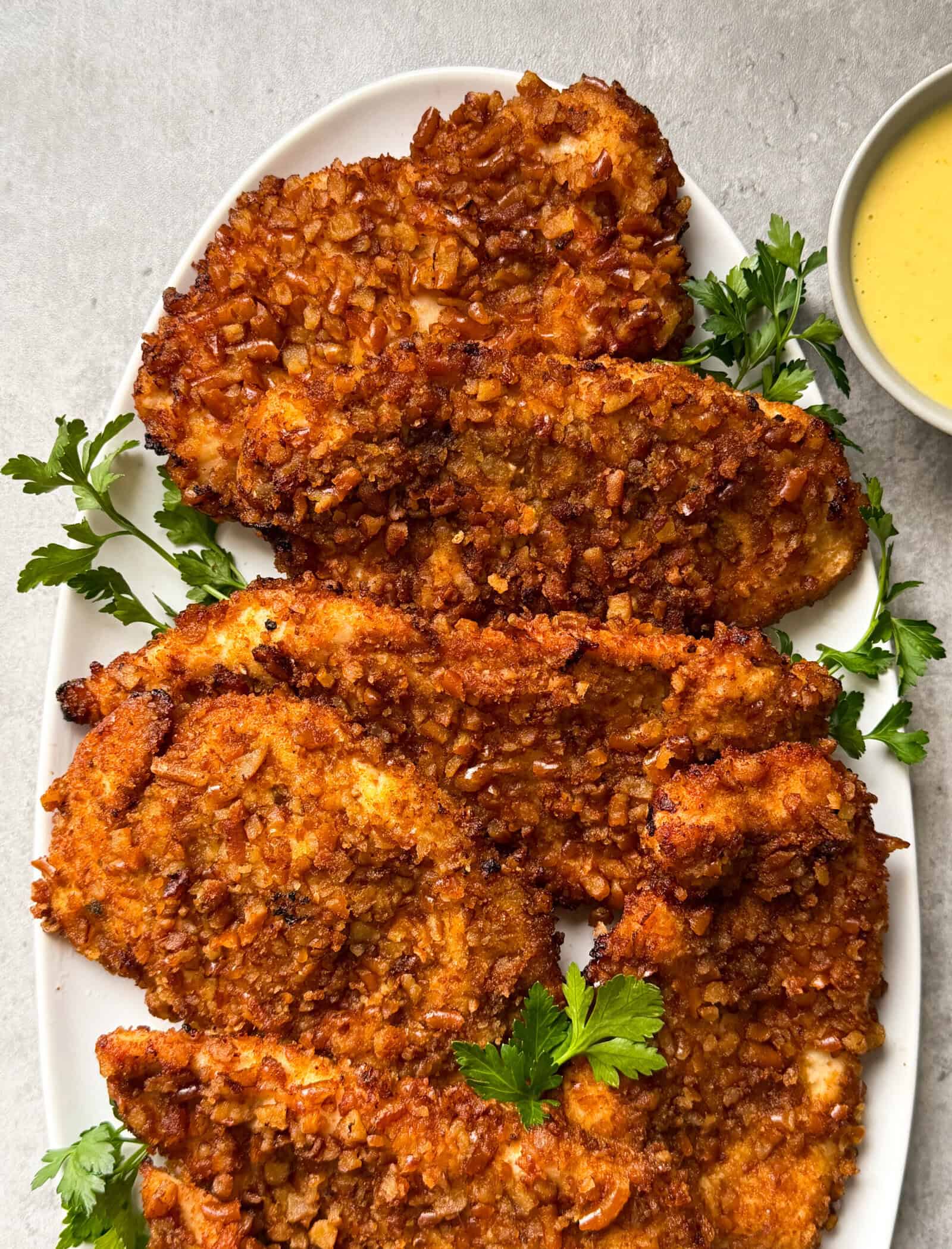 A white oval plate holds five crispy, golden-brown pretzel crusted chicken cutlets garnished with fresh parsley, with a small bowl of yellow dipping sauce beside them—an irresistible pretzel crusted chicken recipe.
