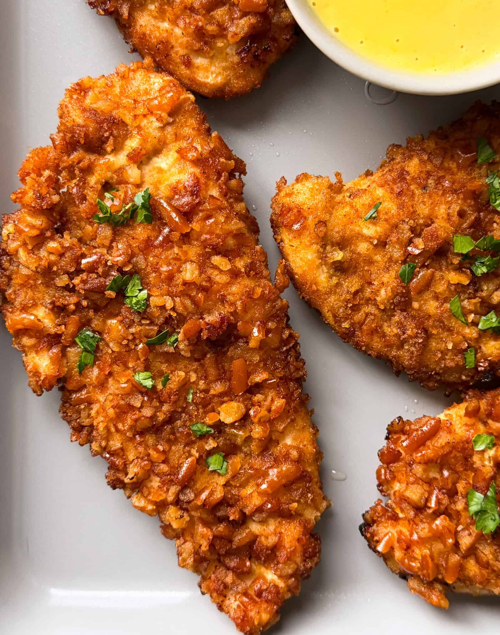 Close-up of crispy, golden-brown pretzel crusted chicken tenders garnished with chopped parsley on a gray plate, with a small bowl of yellow dipping sauce in the corner.