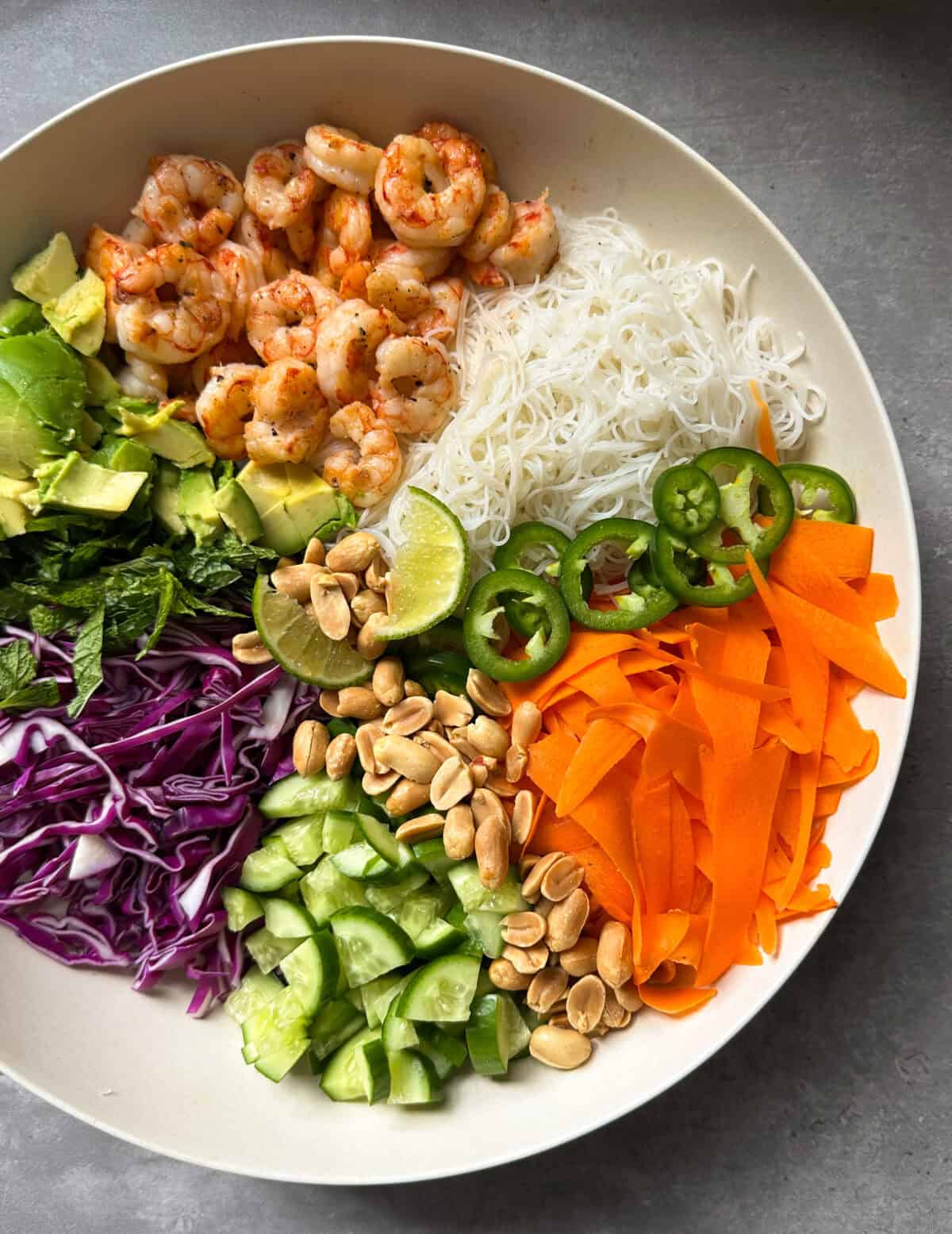A bowl filled with shrimp, vermicelli noodle salad, vermicelli noodles, sliced avocado, mint, shredded purple cabbage, cucumber, carrots, lime wedges, jalape&ntilde;o slices, and peanuts arranged in colorful sections&mdash;like a vibrant spring roll salad on a gray surface.