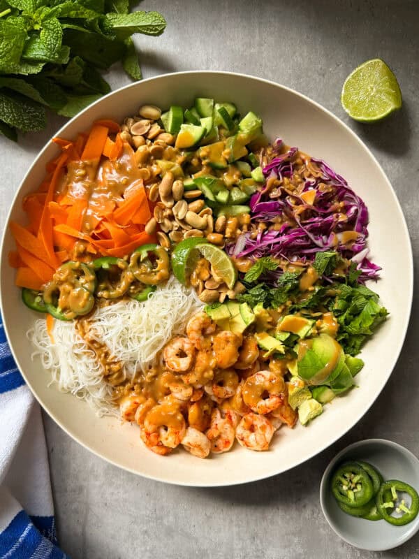 A vibrant spring roll salad bowl filled with shrimp, rice noodles, shredded carrot, cucumber, cabbage, avocado, peanuts, cilantro, jalapeño slices, and peanut sauce—garnished with lime. Mint and a cut lime rest nearby on a gray surface.
