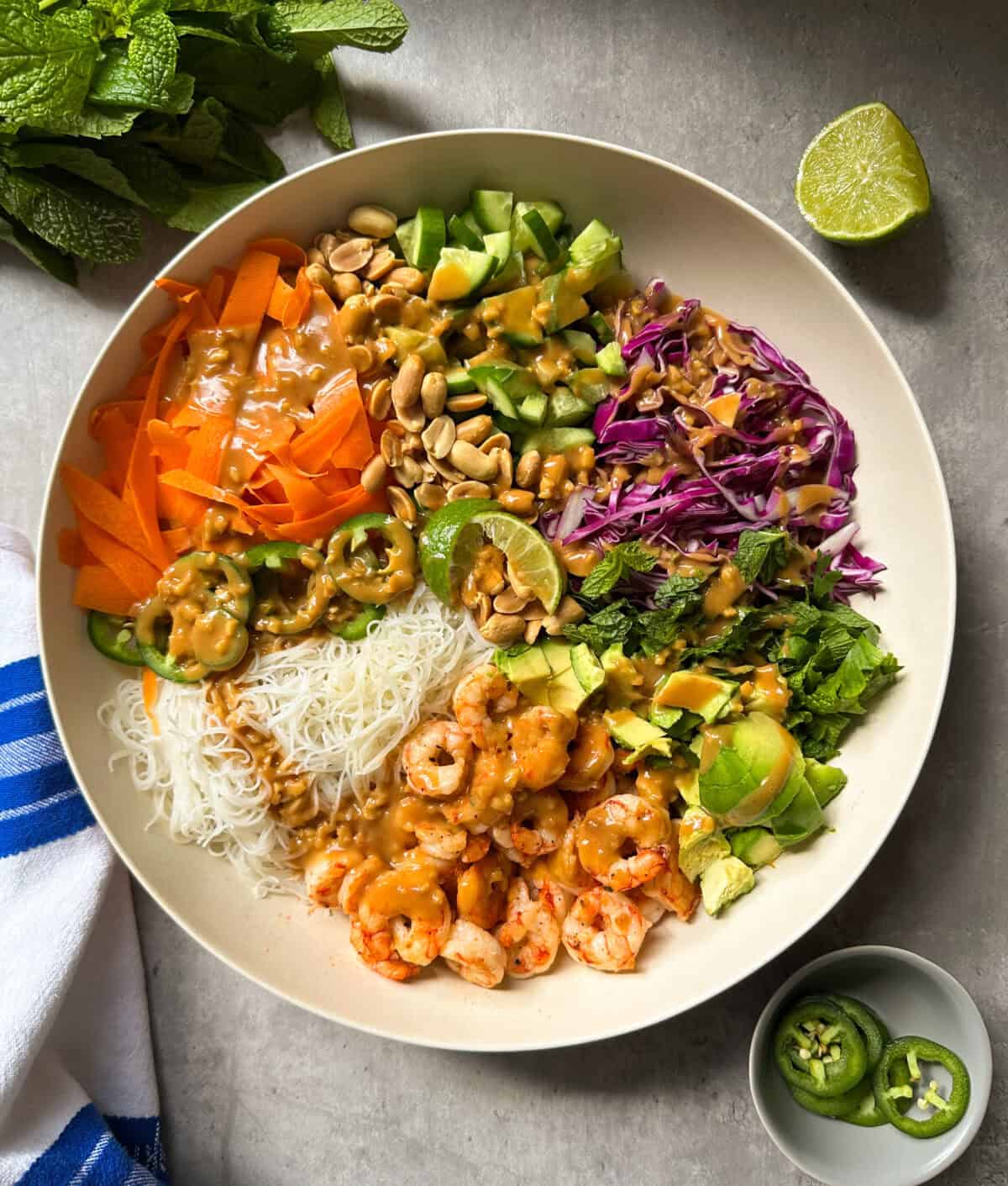 A vibrant spring roll salad bowl filled with rice noodles, vermicelli noodle salad, carrots, cucumber, red cabbage, cilantro, jalape&ntilde;os, avocado, peanuts, and peanut sauce. A lime wedge and fresh mint sit beside the bowl.