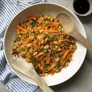 A large bowl of stir-fried noodles with sliced carrots, green beans, and chicken pieces—just like a classic chicken chow mein recipe—topped with sesame seeds. Two wooden utensils rest in the bowl. A small dish of soy sauce sits beside a blue-and-white cloth.