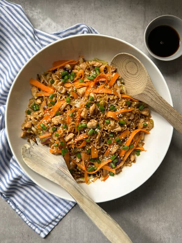 A large bowl of stir-fried noodles with sliced carrots, green beans, and chicken pieces—just like a classic chicken chow mein recipe—topped with sesame seeds. Two wooden utensils rest in the bowl. A small dish of soy sauce sits beside a blue-and-white cloth.