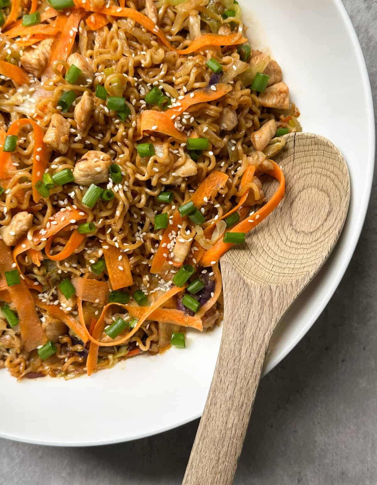 A white plate filled with chicken chow mein—stir-fried noodles, shredded carrots, chicken pieces, green onions, and sesame seeds—with a wooden spoon resting on the edge, showcasing a classic chicken chow mein recipe.