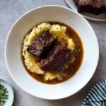 Braised Beef Short Ribs served over creamy mashed potatoes with rich gravy, garnished with chopped herbs, in a white bowl on a gray surface. A striped towel and additional garnishes are nearby, making this Braised Beef Short Ribs Recipe perfect for cozy gatherings.