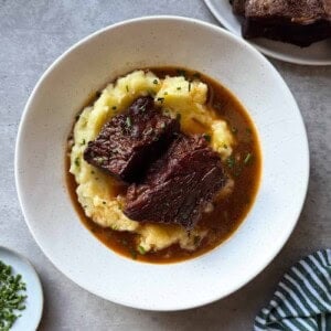 Braised Beef Short Ribs served over creamy mashed potatoes with rich gravy, garnished with chopped herbs, in a white bowl on a gray surface. A striped towel and additional garnishes are nearby, making this Braised Beef Short Ribs Recipe perfect for cozy gatherings.