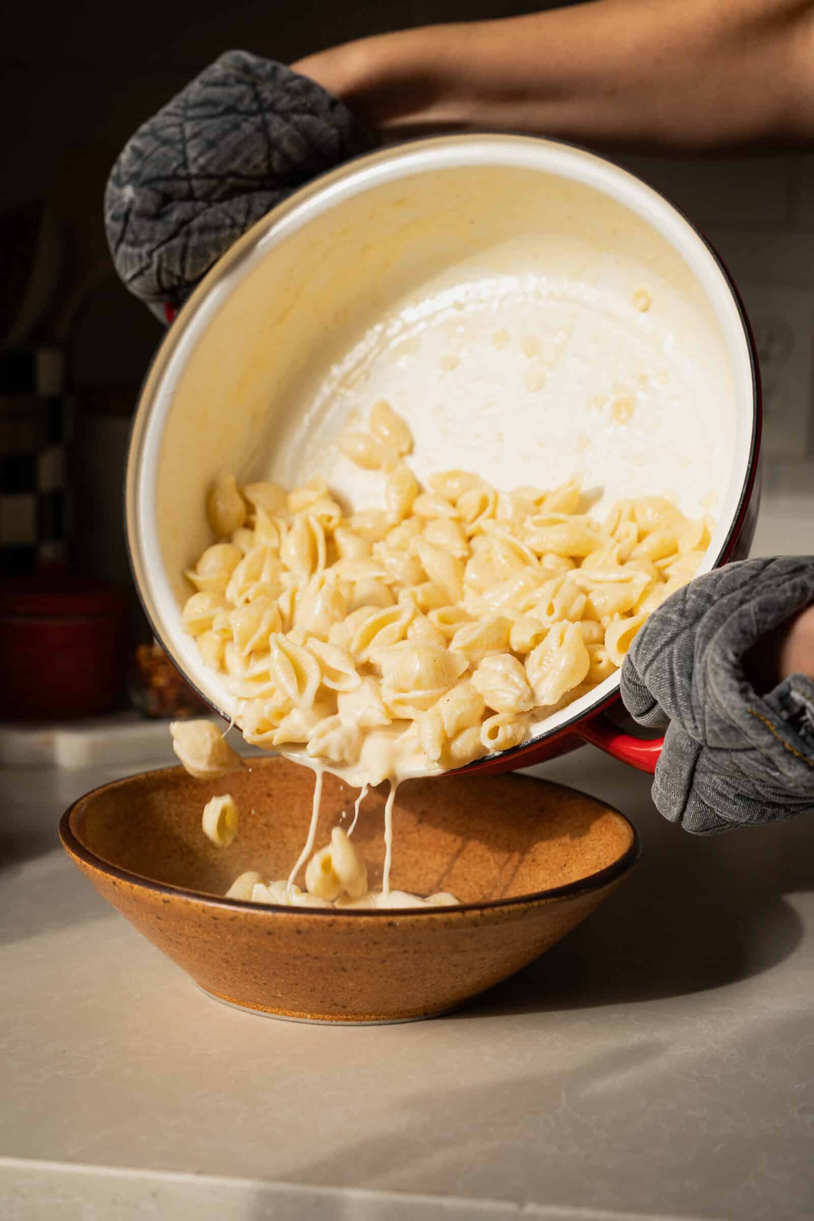 A person wearing gray oven mitts pours creamy lemon pasta from a white pot into a brown bowl on a kitchen counter. The melted cheese stretches between the shells and the pot, showcasing this delicious creamy lemon pasta recipe.