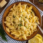 A bowl of creamy lemon pasta topped with grated cheese and chopped herbs sits on a blue-striped napkin, with lemon slices, fresh dill, a wedge of cheese, and a glass of red wine nearby.
