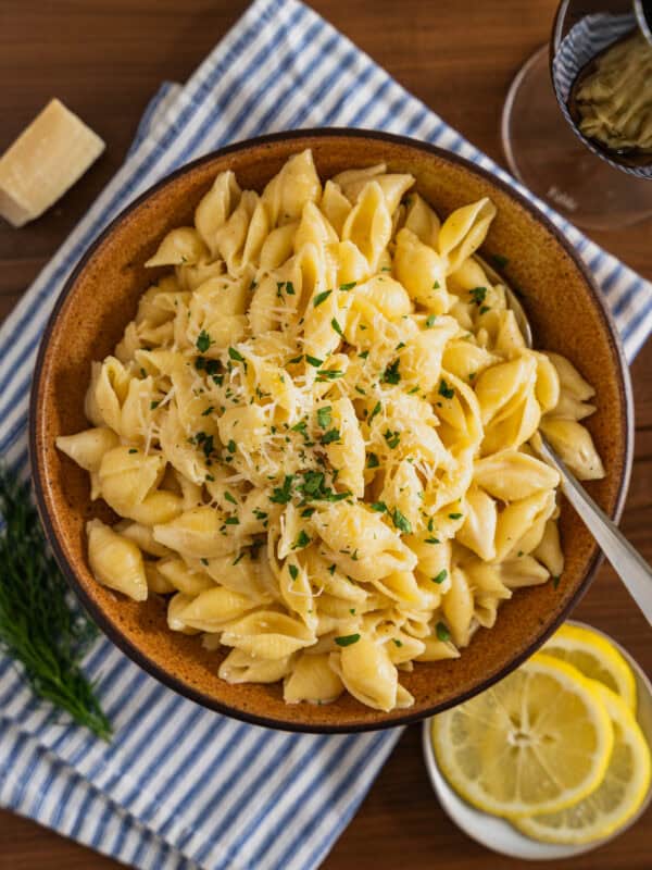 A bowl of creamy lemon pasta topped with grated cheese and chopped herbs sits on a blue-striped napkin, with lemon slices, fresh dill, a wedge of cheese, and a glass of red wine nearby.