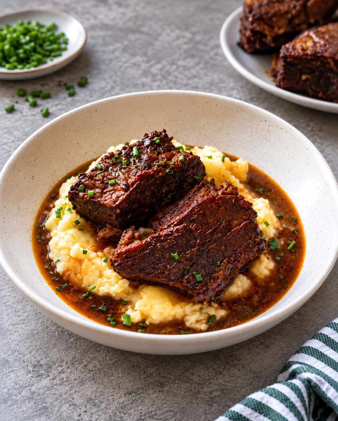A bowl of braised beef short ribs, inspired by a classic Braised Beef Short Ribs Recipe, served over creamy mashed potatoes, garnished with chopped chives and surrounded by a rich brown sauce.