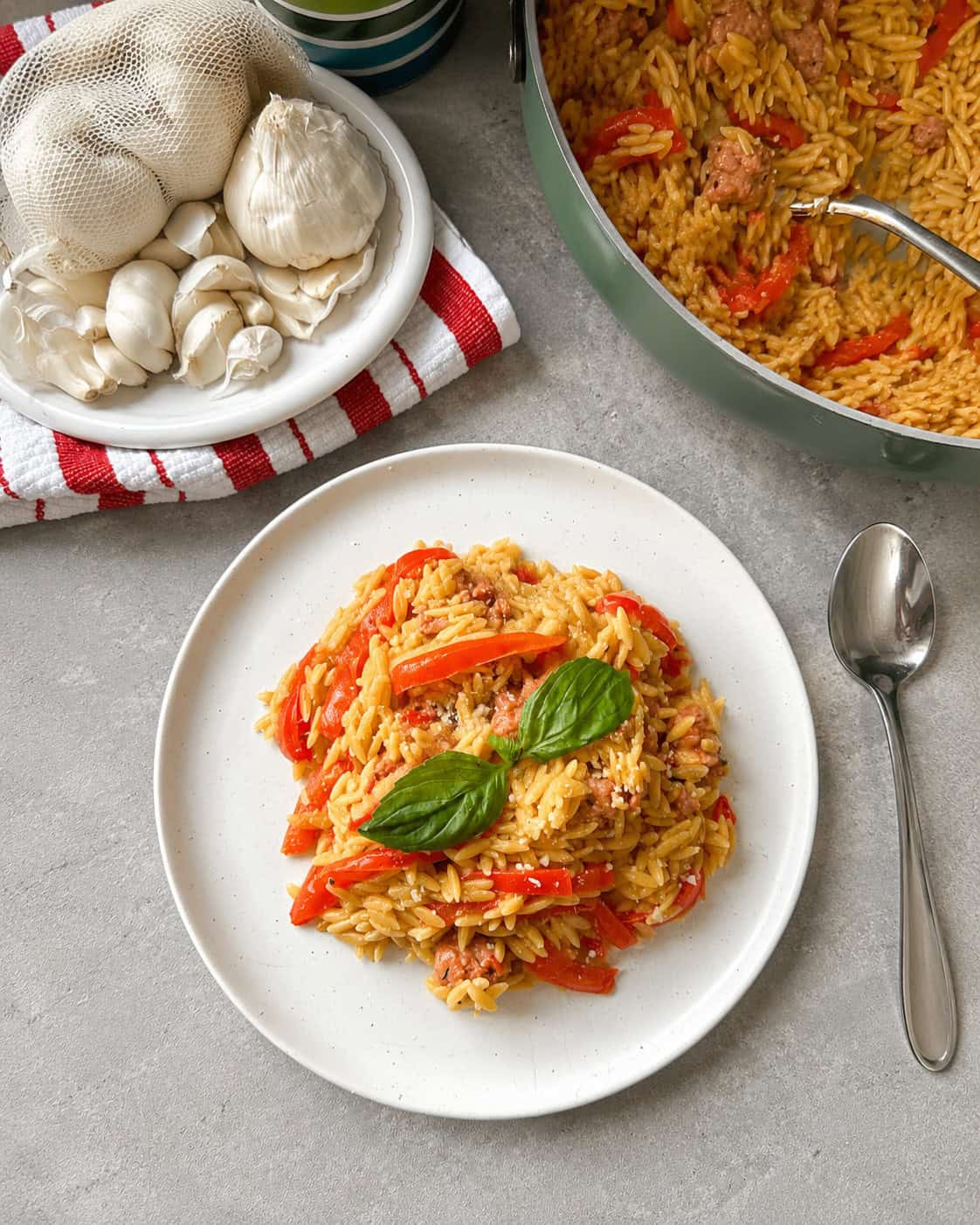 A plate of Fried Sausage and Pepper Orzo features orzo pasta with red bell peppers, herbs, and savory fried sausage, garnished with fresh basil. A skillet of the same dish, a spoon, garlic bulbs, and mushrooms on a red-striped towel are nearby.