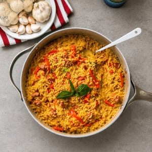 A skillet of yellow rice with fried sausage and red bell peppers, garnished with fresh basil, sits on a gray countertop near a plate of garlic bulbs and a colorful cup.