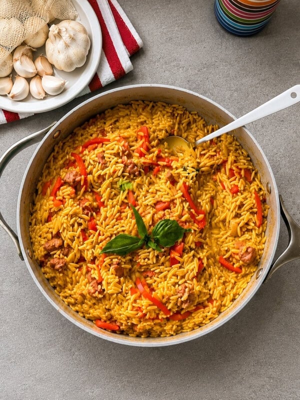 A skillet of yellow rice with fried sausage and red bell peppers, garnished with fresh basil, sits on a gray countertop near a plate of garlic bulbs and a colorful cup.