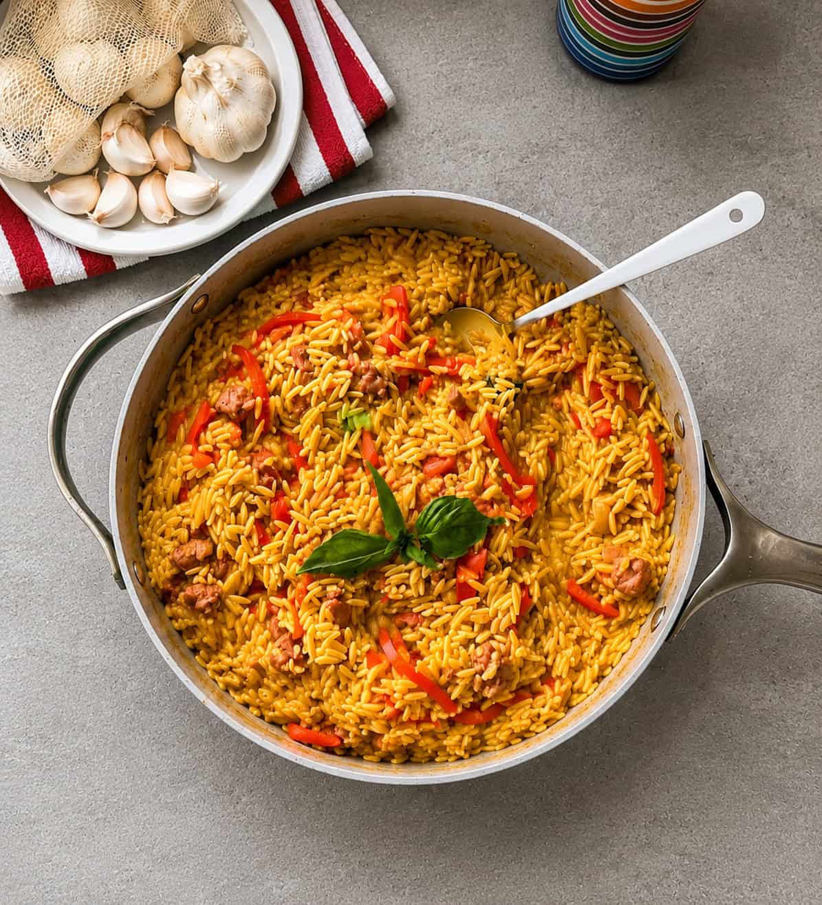 A skillet of yellow rice with fried sausage and red bell peppers, garnished with fresh basil, sits on a gray countertop near a plate of garlic bulbs and a colorful cup.