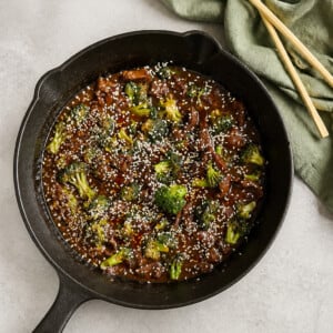 A cast iron skillet filled with beef and broccoli in sauce, topped with sesame seeds. A green cloth and a pair of wooden chopsticks are beside the skillet on a light-colored surface.