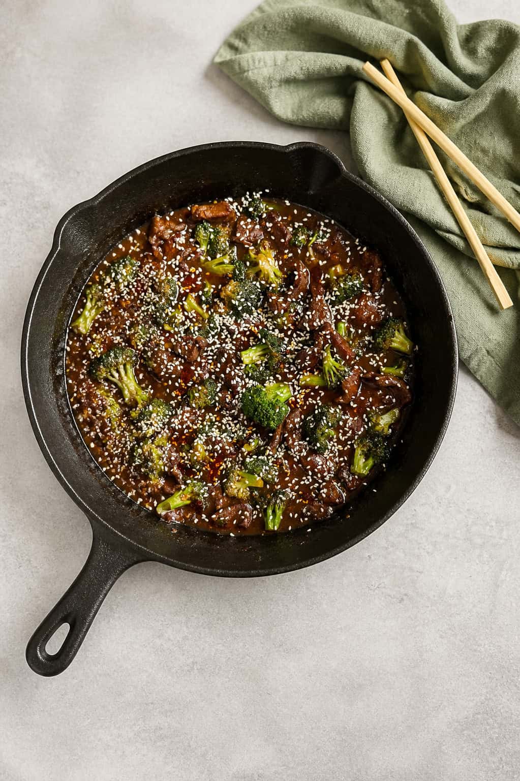 A cast iron skillet filled with beef and broccoli in sauce, topped with sesame seeds. A green cloth and a pair of wooden chopsticks are beside the skillet on a light-colored surface.
