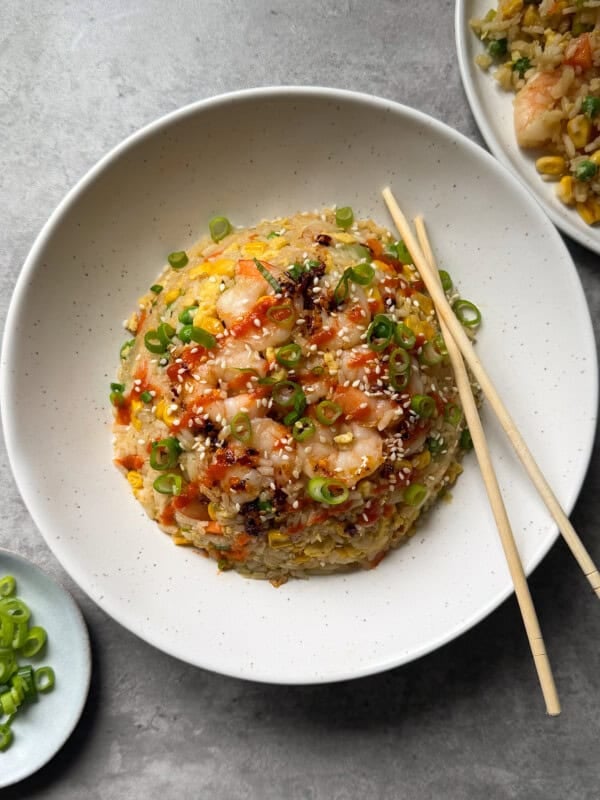 A bowl of Shrimp Fried Rice topped with chopped green onions, sesame seeds, and chili sauce, served with chopsticks. A small plate of green onions and another plate of fried rice are nearby—delicious inspiration for your next Shrimp Fried Rice Recipe.