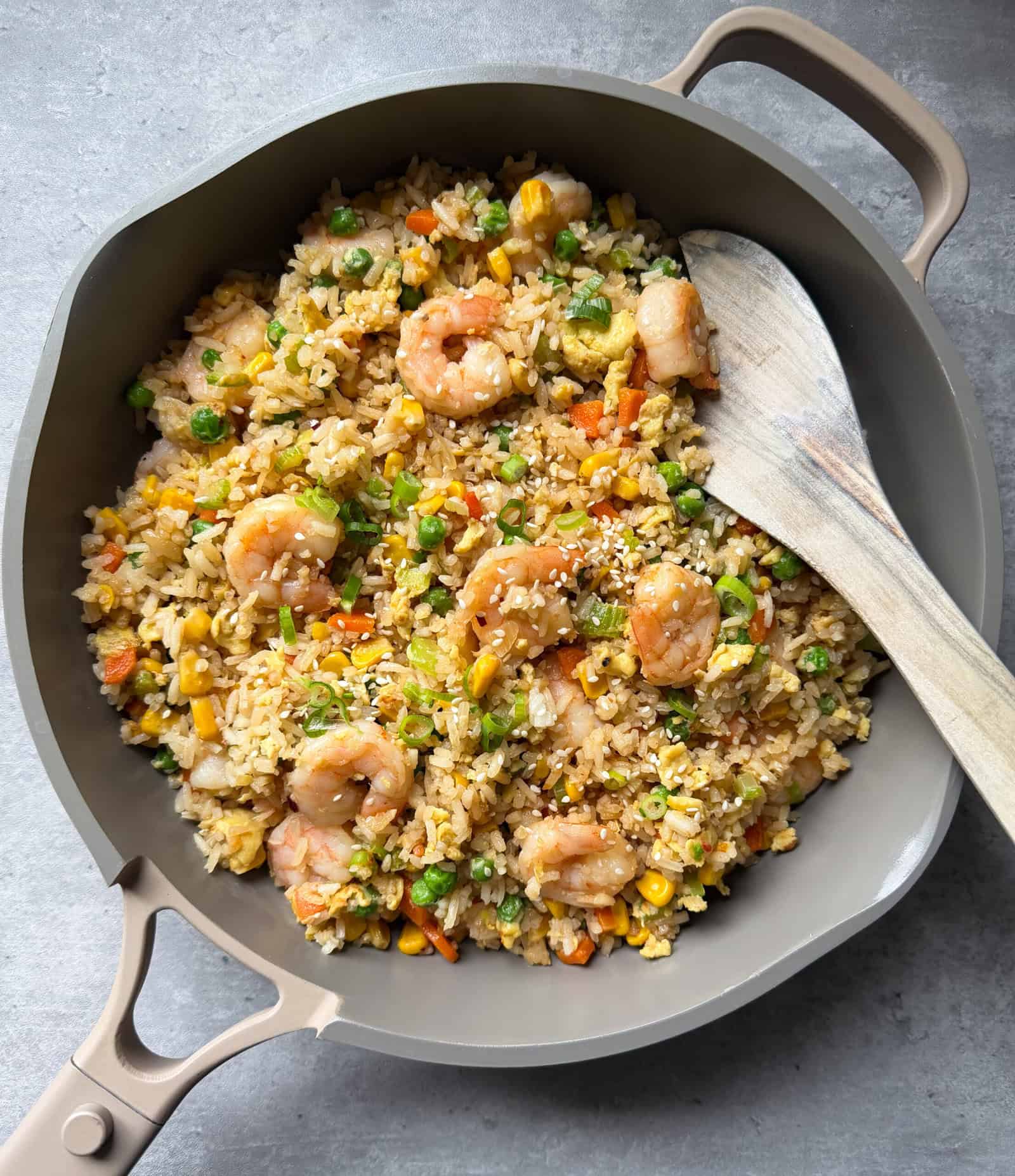 A beige skillet filled with Shrimp Fried Rice—shrimp, peas, carrots, and corn—is shown on a grey surface. A wooden spatula rests on the skillet’s edge, ready to help you master this easy Shrimp Fried Rice recipe.
