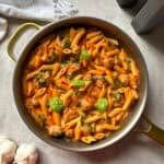 A pan of Spicy Penne Rosé in a creamy tomato sauce with sausage, spinach, and fresh basil sits on a gray surface near garlic bulbs, salt and pepper shakers, and a white cloth.