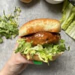 A hand holding a fried chicken sandwich with lettuce on a bun. Shredded lettuce and a knife with a bowl of mayonnaise are visible in the background on a gray surface.
