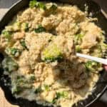 A skillet filled with creamy risotto, featuring pieces of chicken and bright green broccoli. A spoon is lifting a serving, highlighting the creamy texture and well-blended ingredients.