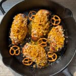 Four breaded chicken breasts garnished with crushed pretzels and drizzled with sauce are cooking in a black skillet. Whole pretzels are scattered around, adding a decorative touch. The skillet is set on a wooden surface.