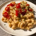 A plate of creamy macaroni and cheese topped with halved cherry tomatoes and sprinkled with chopped green herbs on a stone-textured surface.