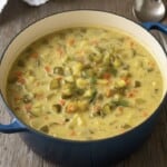 A blue pot filled with creamy Zupa Ogórkowa, or Dill Pickle Soup, containing diced pickles, carrots, and potatoes, garnished with fresh dill, sits on a wooden table next to a spoon.