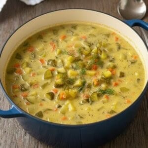 A blue pot filled with creamy Zupa Ogórkowa, or Dill Pickle Soup, containing diced pickles, carrots, and potatoes, garnished with fresh dill, sits on a wooden table next to a spoon.