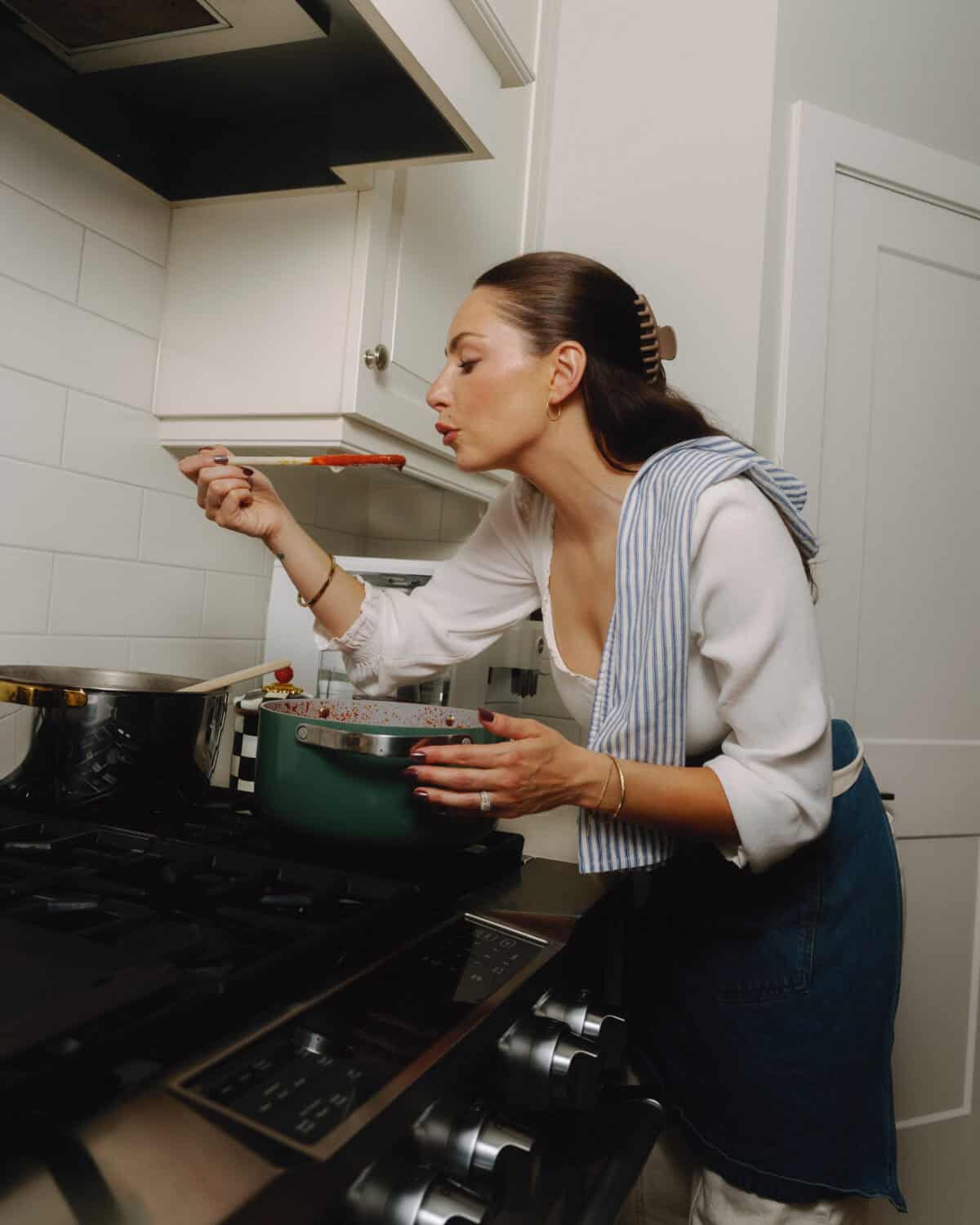 A woman in a kitchen tastes her homemade tomato sauce from a spoon over the stove, holding a pot. She wears a white top, blue apron, and has a striped towel over her shoulder—classic sunday sauce vibes.