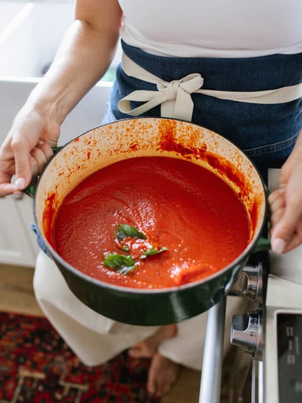 A person wearing a white top and apron holds a green pot filled with rich Homemade Tomato Sauce, garnished with fresh basil leaves, in a cozy kitchen setting—a perfect scene for preparing your favorite sunday sauce.