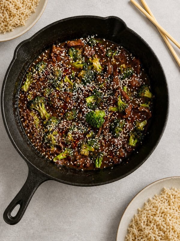 A cast iron skillet filled with beef and broccoli stir-fry, topped with sesame seeds. Plates of cooked rice and a pair of chopsticks are nearby on a light surface.