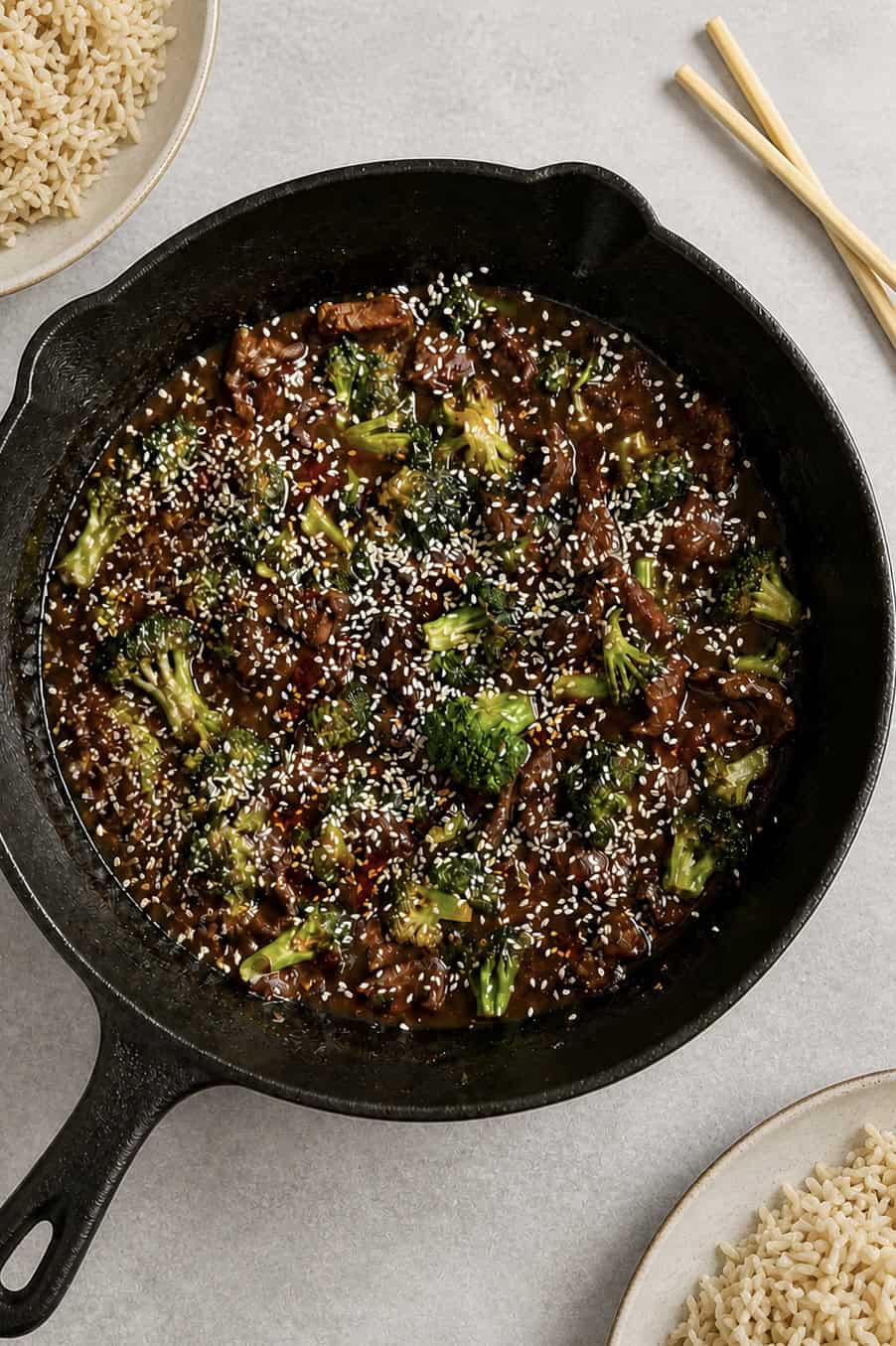 A cast iron skillet filled with beef and broccoli in a dark sauce, topped with white sesame seeds. Plates of brown rice and a pair of wooden chopsticks are visible nearby on a light surface.